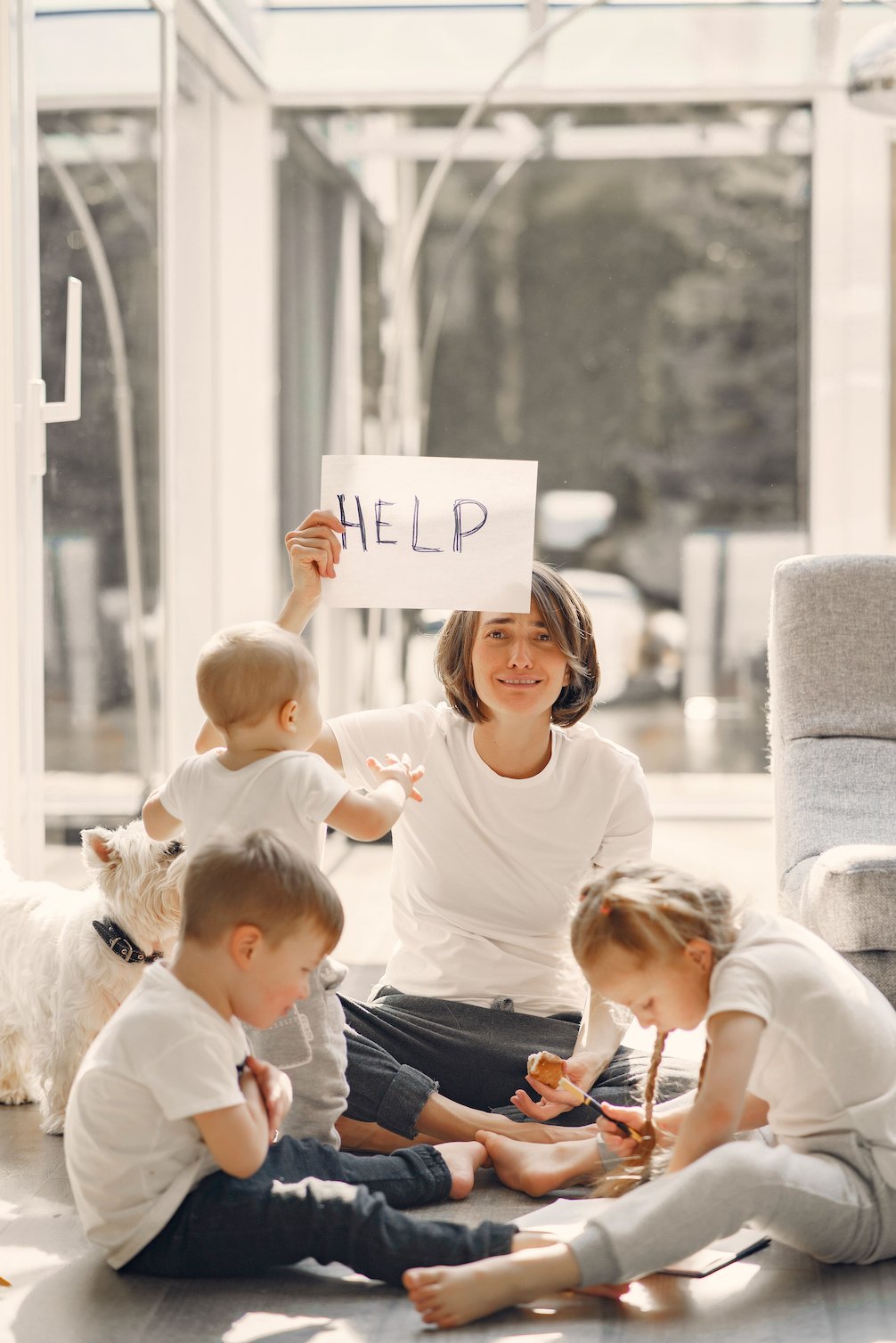 An overwhelmed mom holding a HELP sign while her three young children play around her — maternal mental load and depletion